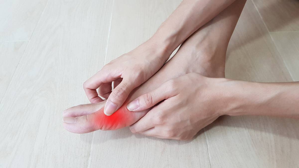 A person grimaces in pain while sitting on a wooden floor, holding their injured foot.