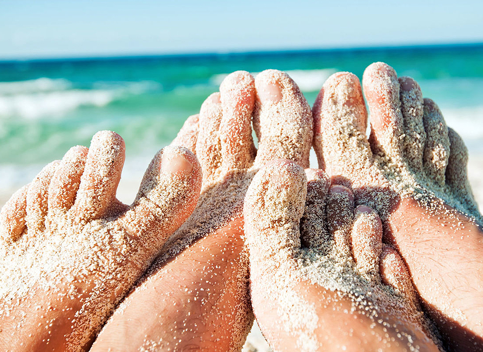 Two pairs of feet resting in soft sand on a beach, with gentle waves lapping in the background.